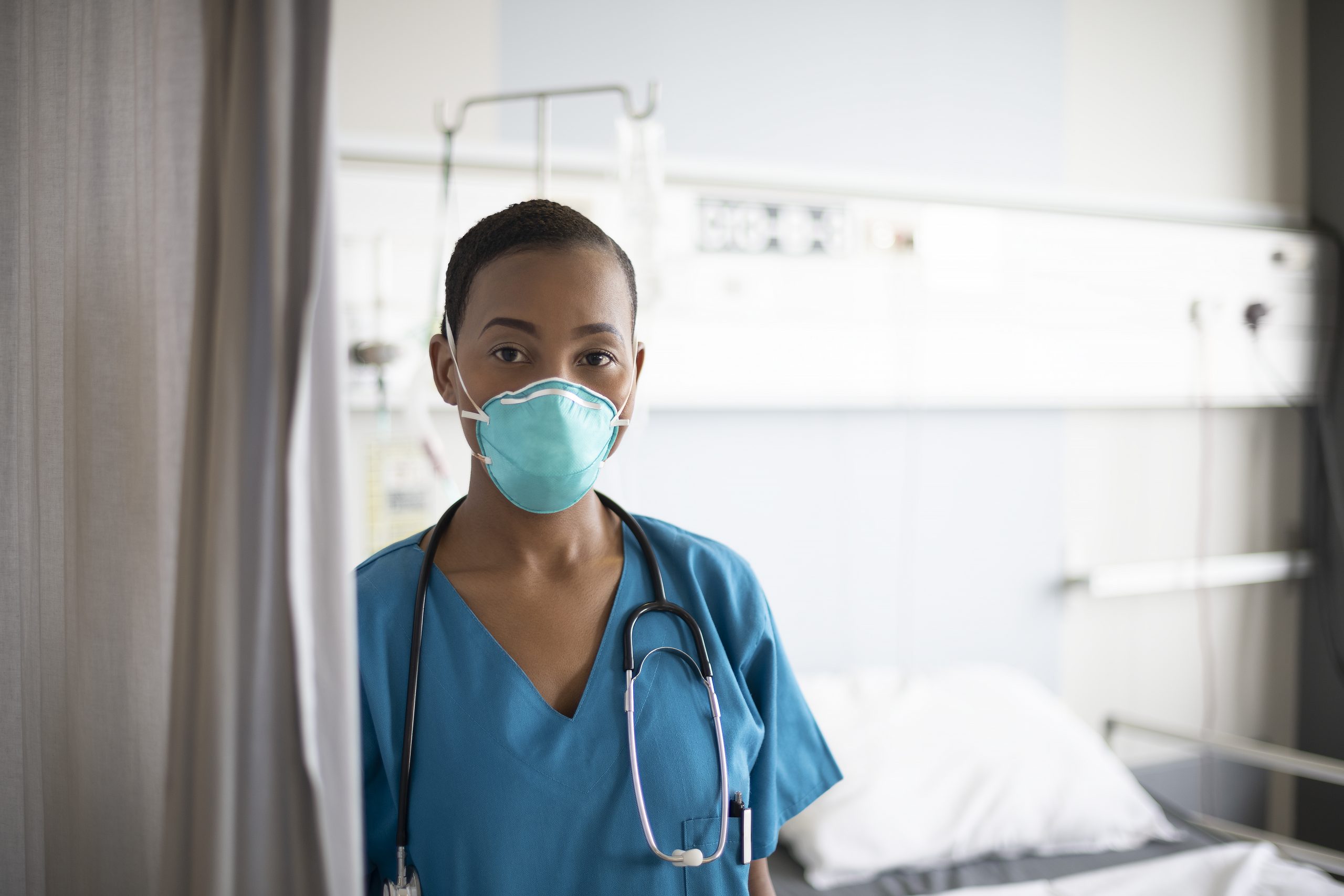 Close-up of African female nurse in mid 20s wearing blue scrubs, N95 face mask, and stethoscope around her neck standing in hospital room with empty bed