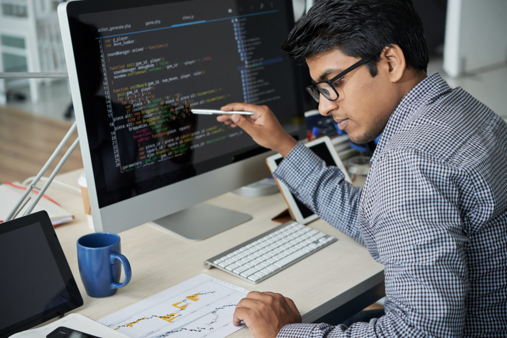 Indian programmer in eyeglasses pointing at computer monitor and checking the statistics of website in document while working at office