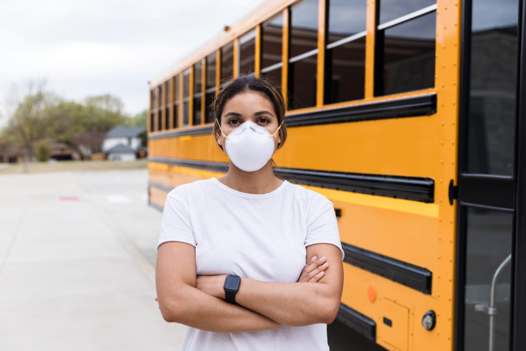 Female school bus driver, wearing an N95 mask, stands next to a school bus