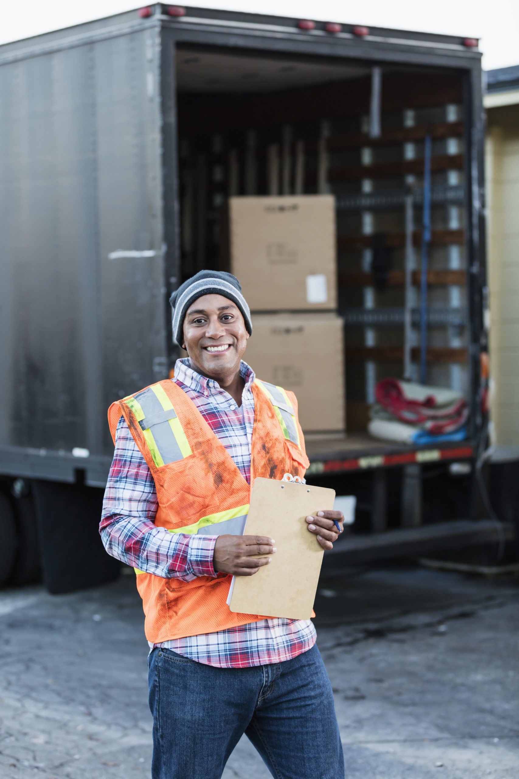 Mature African-American man in wearing a reflective vest, holding a clipboard, standing in front of a delivery truck