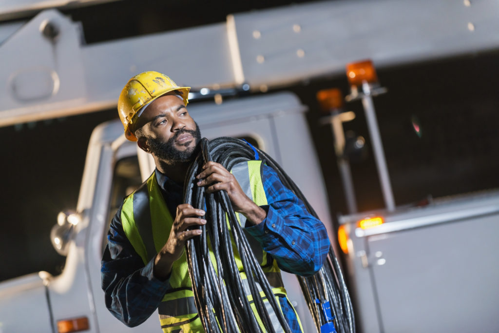African American man wearing a hard hat and safety vest, standing in front of a bucket truck or cherry picker, carrying cables on his shoulder