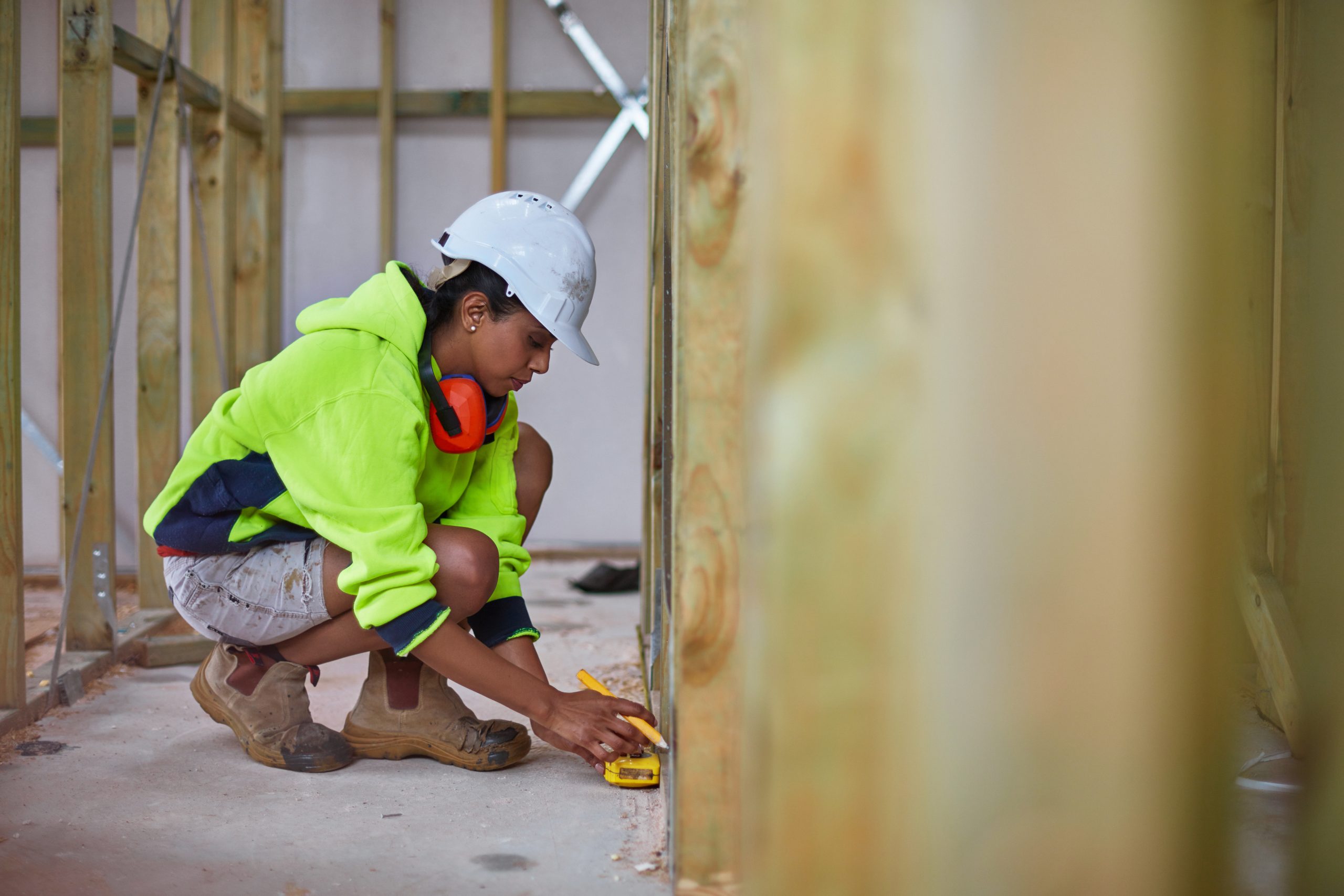 Full length of construction worker using tape measure while working. Female engineer is in reflective clothing. She is crouching on floor at construction site.