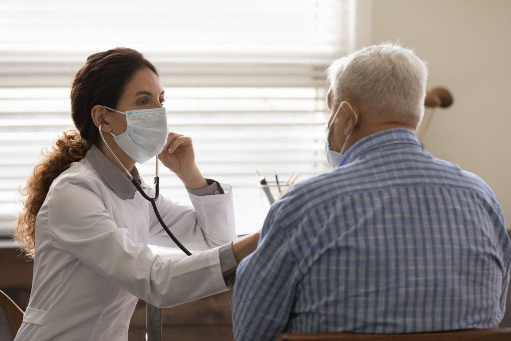 Confident female doctor wearing face mask checking mature man lungs, using stethoscope, listening to elderly patient heartbeat, breath, elderly generation healthcare, medical checkup concept
